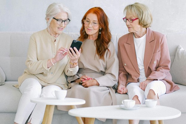 Three elderly women sit on a couch looking at a smartphone.