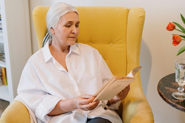 An elderly woman sitting in a yellow armchair, reading a book.